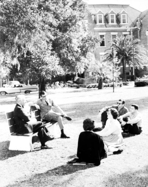 1960s black and white photograph showing a group of male and female students sitting on the ground outside with 2 male professors on a bench