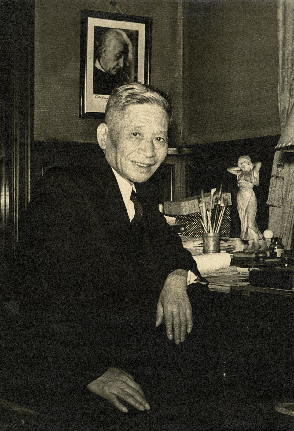 Portrait of Sanehiko Yamamoto sitting at his office desk which is covered with writing materials with the portrait of Einstein behind him. 
